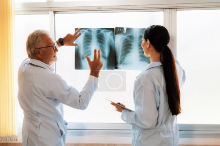 In a hospital sterile room, two professional radiographers hold and examine a radiograph for medical xray diagnosis. Novice doctor seeks advice on a patients condition from experienced older doctor.