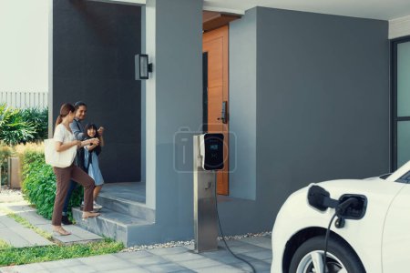 Foto de Progressive young parents and daughter with electric vehicle and home charging station. Green and clean energy from electric vehicles for healthy environment. Eco power from renewable source at home. - Imagen libre de derechos