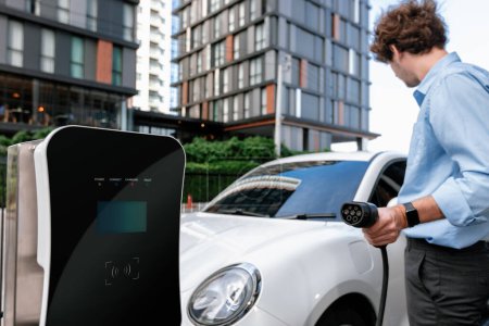 Téléchargez les photos : Progressive businessman insert charger plug from charging station to his electric vehicle with apartment condo building in background. Eco friendly rechargeable car powered by sustainable energy. - en image libre de droit