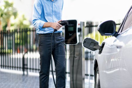 Closeup progressive suit-clad businessman with his electric vehicle recharge his car on public charging station in modern city with power cable plug and renewable energy-powered electric vehicle.
