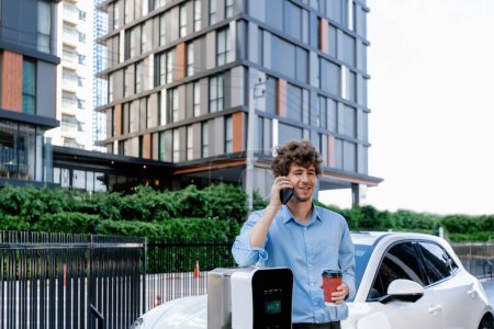 Progressive businessman talking on the phone with coffee, leaning on electric car recharging with public EV charging station, apartment condo residential building on background as green city lifestyle