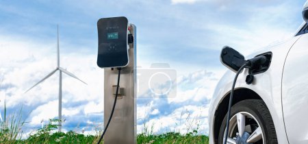 Progressive combination of wind turbine and EV car, future energy infrastructure. Electric vehicle being charged at charging station powered by renewable energy from wind turbine in the countryside.