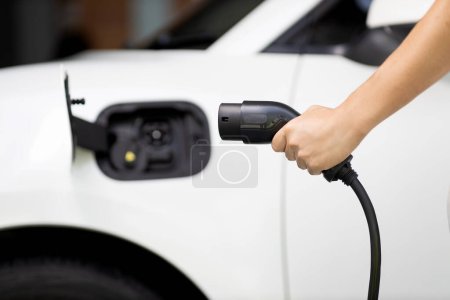 Foto de Focus hand holding EV charger plug with blurred background of progressive electric vehicle and socket parking in home garage with electric charging station powered by clean and sustainable energy. - Imagen libre de derechos