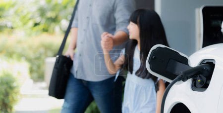 Photo for Focus electric car recharging at home charging station with blurred father and daughter walking in background. Progressive green and clean energy vehicle for healthy environment lifestyle concept. - Royalty Free Image
