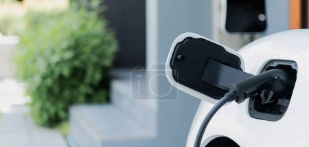 Foto de Closeup electric vehicle plugged-in with cable from charging point powered for progressive concept by alternative clean energy rechargeable EV car at home charging station. - Imagen libre de derechos