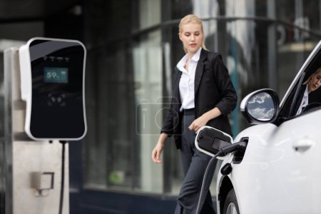 Focus public EV charging station for electric car with blurred progressive businesswoman walking in background. Electric car driven by clean and sustainable energy for ecological concern.
