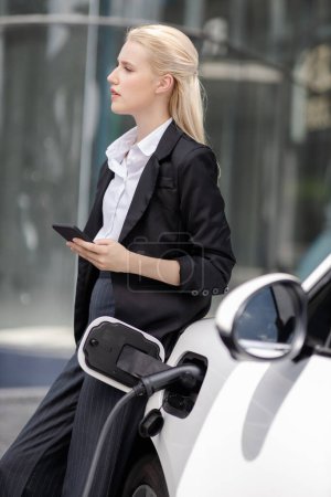 Foto de Businesswoman wearing black suit using smartphone, leaning on electric car recharge battery at charging station in city residential building with condos and apartment. Progressive lifestyle concept. - Imagen libre de derechos