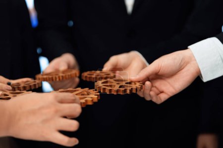 Closeup hand holding wooden gear by businesspeople wearing suit for harmony synergy in office workplace concept. Group of people hand making chain of gears into collective form for unity symbol.