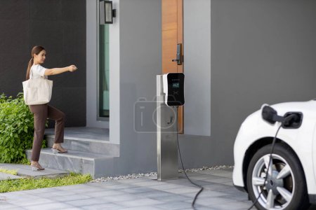 Foto de Asian woman lock electric vehicle with remote car key at her garage, EV car recharge battery at home charging station. Alternative clean energy applied in daily life as progressive lifestyle concept. - Imagen libre de derechos