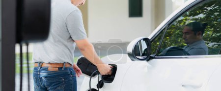 Foto de Progressive asian man install cable plug to his electric car with home charging station in the backyard. Concept use of electric vehicles in a progressive lifestyle contributes to clean environment. - Imagen libre de derechos