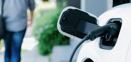 Foto de Focus electric car charging at home charging station with blurred progressive man walking in the background. Electric car using renewable clean for eco-friendly concept. - Imagen libre de derechos