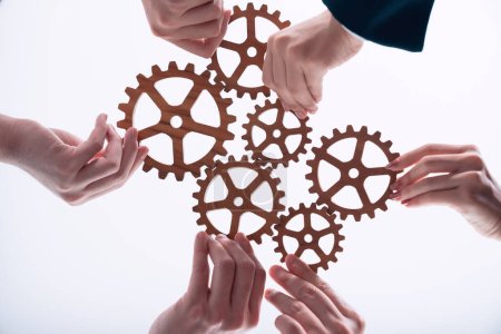 Foto de Hand holding wooden gear by businesspeople wearing suit for harmony synergy in office workplace concept. Isolated background. Bottom view of people hand make chain of gear into collective unity symbol - Imagen libre de derechos