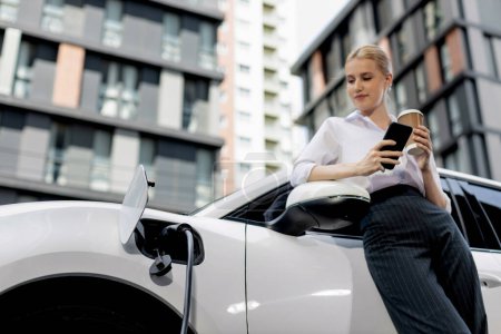 Focus businessman using phone, leaning on electric vehicle, holding coffee with blurred city residential condo buildings in background as progressive lifestyle by renewable and sustainable EV car.