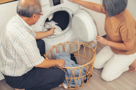 Téléchargez les photos : Senior couple working together to complete their household chores at the washing machine in a happy and contented manner. Husband and wife doing the usual tasks in the house. - en image libre de droit