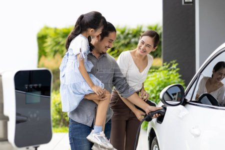 Foto de Progressive young parent teach daughter how to recharge or refuel EV car at home charging station. Green and clean energy from electric vehicle for healthy environment. Eco power from renewable source - Imagen libre de derechos