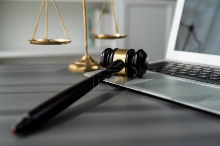 Symbolisch für Gerechtigkeit und rechtliche Autorität, goldene Waage und Hammer auf dem Schreibtisch mit Laptop im Hintergrund der Kanzlei, die das Konzept der Gleichheit und des fairen Urteils von Anwälten und Richtern widerspiegeln. Gleichgewicht