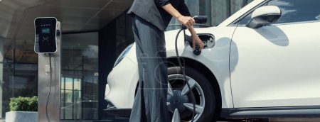 Closeup progressive suit-clad businesswoman with her electric vehicle recharge her car on public charging station in modern city with power cable plug and renewable energy-powered electric vehicle.