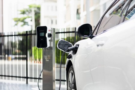 Foto de Focus closeup electric vehicle plugged in with EV charger device from blurred background of public charging station powered by renewable clean energy for progressive eco-friendly car concept. - Imagen libre de derechos