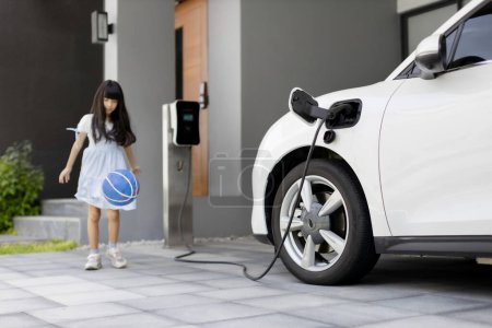 Foto de A playful and happy girl playing around at her home charging station providing a sustainable power source for electric vehicles. Alternative energy for progressive lifestyle. - Imagen libre de derechos
