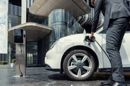 Photo for Below view closeup image of progressive black suit businessman recharge battery of his electric vehicle from public charging station. Renewable and alternative energy powered car concept. - Royalty Free Image