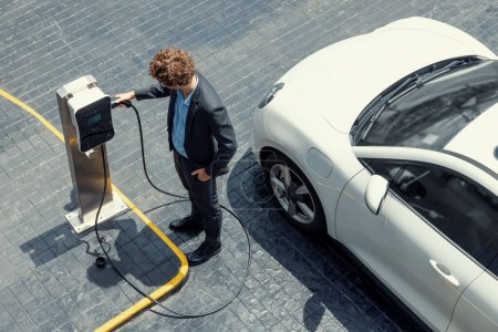 Photo for Aerial view of progressive businessman in black formal suit with his electric vehicle recharging battery at public car park charging station as vehicle powered by sustainable energy concept. - Royalty Free Image