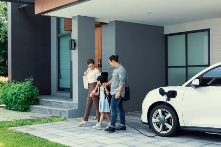 Foto de Progressive young parents and daughter with electric vehicle and home charging station. Green and clean energy from electric vehicles for healthy environment. Eco power from renewable source at home. - Imagen libre de derechos
