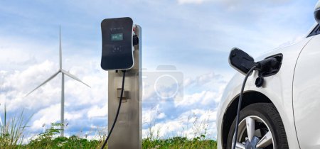 Progressive combination of wind turbine and EV car, future energy infrastructure. Electric vehicle being charged at charging station powered by renewable energy from wind turbine in the countryside.
