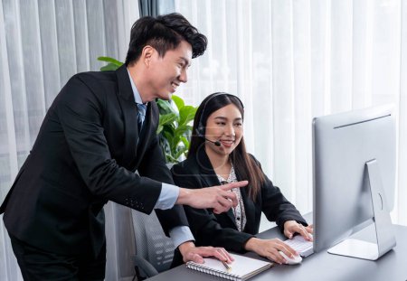 Managerin hilft Callcenter-Betreiberin bei Kundenbetreuung. Ein asiatisches Callcenter mit Kopfhörer und Mikrofon wird im modernen Büro angeleitet. Effiziente Teamarbeit für den Telesaleservice. Jubelnd