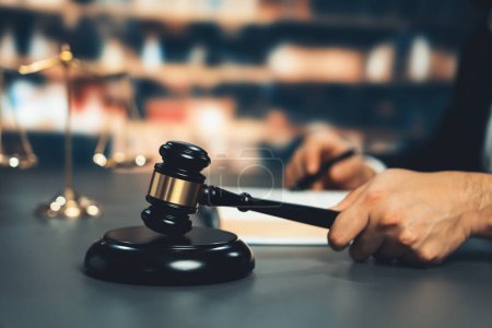 Photo for Focus closeup hand holding black wooden gavel and bang on sounding block by blurred judge in library or courtroom background as concept of litigation justice of judicial system. Equilibrium - Royalty Free Image