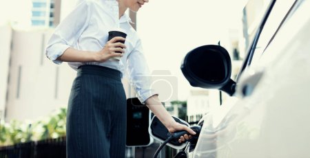 Closeup businesswoman holding coffee, insert EV charger to electric vehicle at public charging station. Eco-friendly car using clean and renewable energy for progressive lifestyle in city.