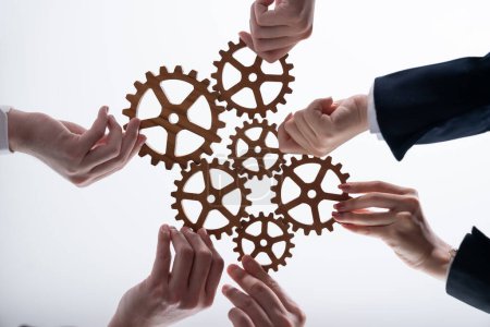 Foto de Hand holding wooden gear by businesspeople wearing suit for harmony synergy in office workplace concept. Isolated background. Bottom view of people hand make chain of gear into collective unity symbol - Imagen libre de derechos