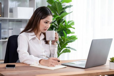 Young asian enthusiastic businesswoman at modern office desk using laptop to work with a cup of coffee. Diligent and attractive office lady working on computer notebook in her office work space.
