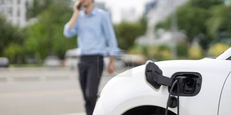Progressive eco-friendly concept of focus parking EV car at public electric-powered charging station in city with blur background of businessman talking on the phone while recharging electric vehicle.