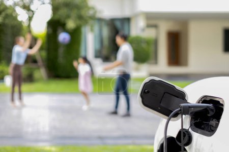 Foto de Focus EV charger connected with EV car at outdoor home charging station with blur progressive family playing together in the background. EV car for alternative clean energy concept. - Imagen libre de derechos