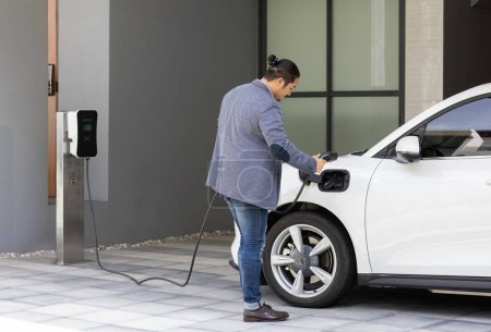 Foto de Progressive asian man install cable plug to his electric car with home charging station in the backyard. Concept use of electric vehicles in a progressive lifestyle contributes to clean environment. - Imagen libre de derechos