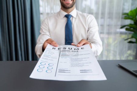Confident man wearing suit in formal office, hand holding resume paper during job interview. Interviewer point of view with candidate handing resume paper in front of camera for consideration. Fervent