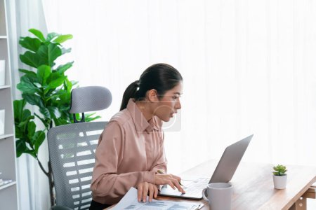 Young asian enthusiastic businesswoman at modern office desk using laptop to work and write notes. Diligent and attractive office lady working on computer notebook in her office work space.