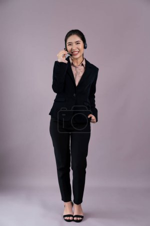 Photo for Attractive full body asian female call center operator with happy face advertises job opportunity on empty space, wearing formal suit and headset on customizable isolated background. Enthusiastic - Royalty Free Image