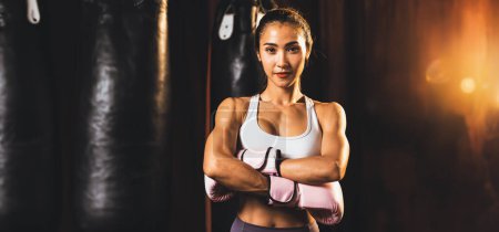 Téléchargez les photos : Femme asiatique Muay boxer thaïlandais poing poing devant la caméra en position de combat prêt à poser à la salle de gym avec équipement de boxe en arrière-plan. Regard de détermination concentré et préparation au défi. Éperon - en image libre de droit