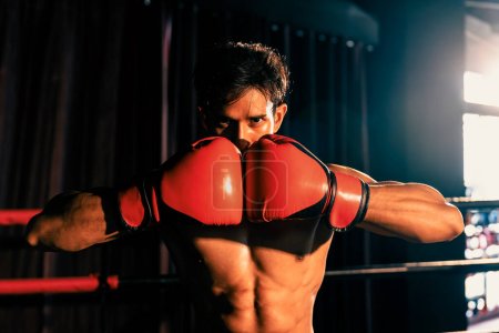 Téléchargez les photos : Muay boxeur thaïlandais punch poisson sa main ensemble devant la caméra dans prêt à se battre position posant à la salle de gym avec équipement de boxe en arrière-plan. Détermination ciblée œil et se préparer pour le défi. Impulsion - en image libre de droit