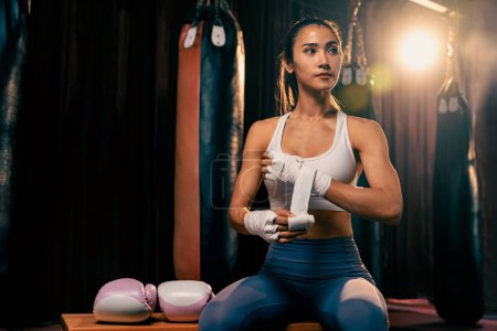 Photo for Determined Asian female Muay Thai boxer with muscularity physical readiness body wraps her hand and dons or wearing boxing glove, preparing for intense boxing training in the ring at gym. Impetus - Royalty Free Image