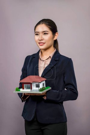 Téléchargez les photos : Jeune femme asiatique portant un costume formel, tenant et mettant en valeur le modèle de la maison sur fond isolé. Idéal pour la publicité de logement, les promotions d'assurance habitation ou l'espace de copie personnalisable. Enthousiaste - en image libre de droit
