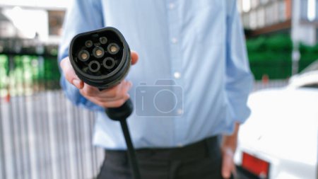 Photo for Suit-clad businessman with progressive ambition leaning pointing power cable plug at camera, charging station at city center. Electric vehicle, EV car driven by renewable energy for green environment. - Royalty Free Image
