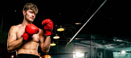 Téléchargez les photos : Combattant de boxe pose torse nu, boxeur homme caucasien portant un gant rouge en position de garde défensive prêt à se battre et frapper à la salle de gym avec sac de coup de pied et équipement de boxe en arrière-plan. Éperon - en image libre de droit