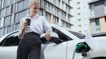 Téléchargez les photos : Femme d'affaires buvant du café après avoir rechargé sa voiture électrique de la station de recharge au parking résidentiel. Futuriste voiture écologique EV utiliser l'énergie alternative dans le mode de vie urbain de la ville. Pertinence - en image libre de droit
