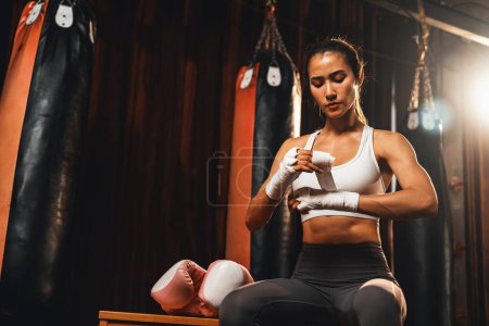 Téléchargez les photos : Femme asiatique déterminée Muay boxeuse thaïlandaise avec muscularité corps de préparation physique enveloppe sa main et dons ou porter un gant de boxe, se préparant à un entraînement intense de boxe sur le ring à la salle de gym. Impulsion - en image libre de droit