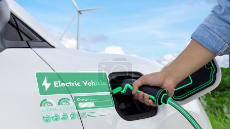 Photo for Hand insert EV charger and recharge electric car from charging station displaying futuristic battery status hologram with wind turbine farm background. Smart sustainable and alternative energy. Peruse - Royalty Free Image