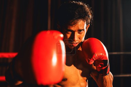 Foto de El boxeador Muay Thai golpea su puño frente a la cámara en posición lista para luchar posando en el gimnasio con equipo de boxeo en segundo plano. Ojos de determinación enfocados y prepararse para el desafío. Impulso - Imagen libre de derechos
