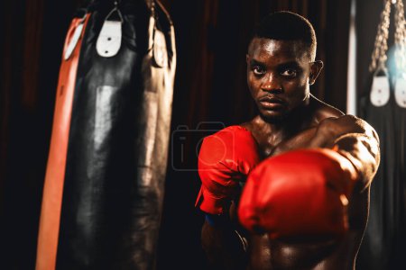 Téléchargez les photos : Combattant de boxe pose torse nu, boxeur noir afro-américain frapper son poing en position agressive et prêt à se battre à la salle de gym avec sac de coups de pied et équipement de boxe en arrière-plan. Impulsion - en image libre de droit