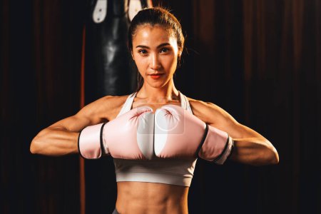 Téléchargez les photos : Femme asiatique Muay boxer thaïlandais frapper son poing et la main ensemble devant la caméra dans prêt à se battre position posant à la salle de gym avec équipement de boxe en arrière-plan. Détermination œil pour défi. Impulsion - en image libre de droit